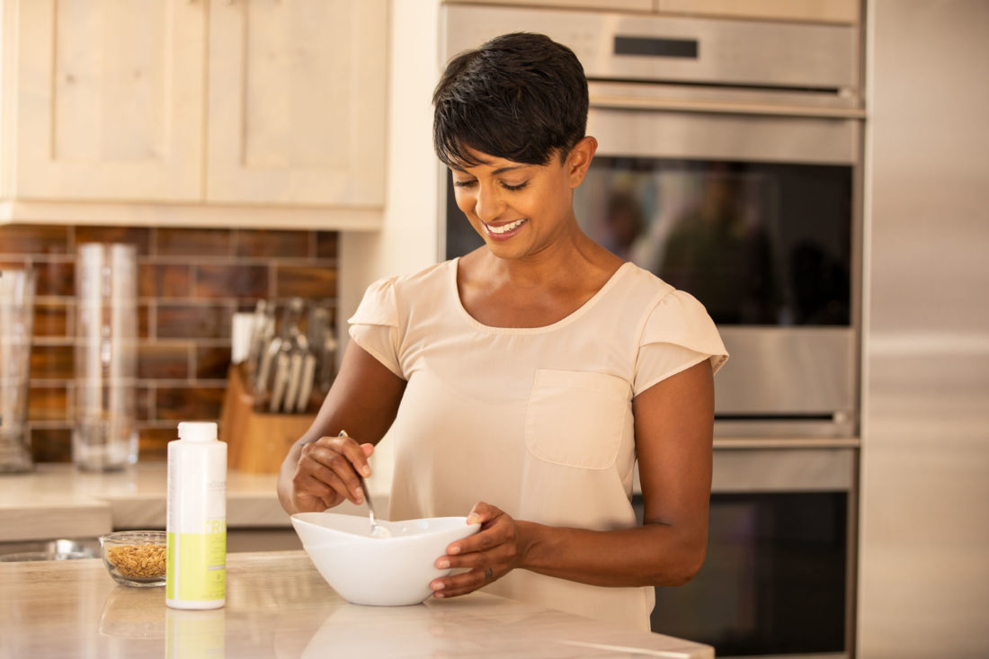 A woman stands in a kitchen, mixing ingredients in a bowl. A bottle of Modere Trim stands on the countertop next to her.