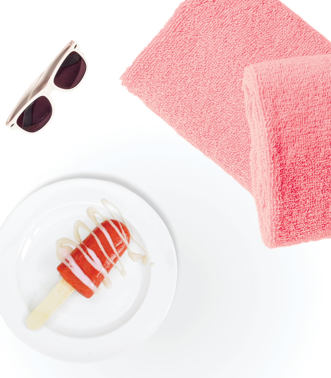 A raspberry lemonade popsicle on a plate. A pair of sunglasses and pink beach towel sit next to the plate.