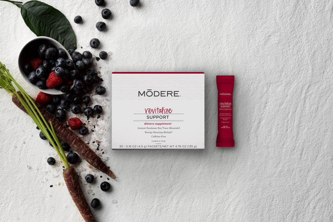 A box of Modere Revitalize. Two dirty carrots and a handful of blueberries and raspberries are scattered to the left of the box. A Revitalize sachet is to the right of the box.