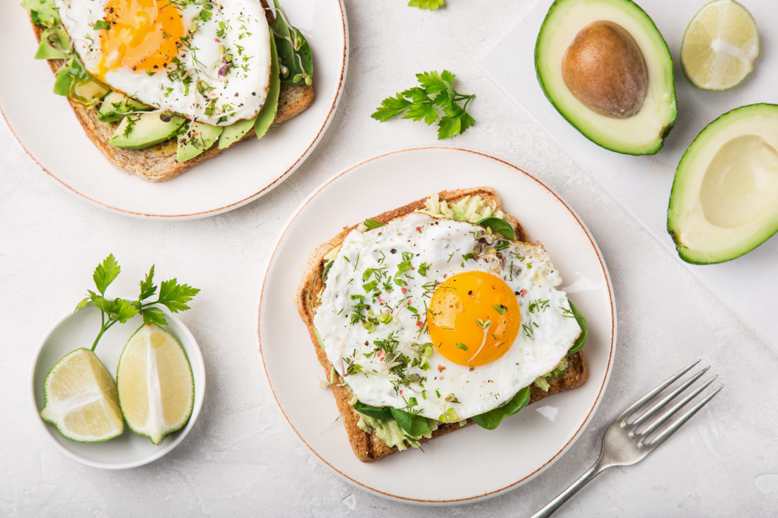 2 plates of Avocado Toast with a fried egg. Lime wedges and avocado halves sit next to the plates in small dishes.