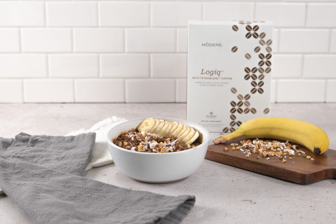 A bowl of Modere Logiq chocolate peanut butter oatmeal, resting on a kitchen counter. To the left of the bowl are two dish towels. Behind the bowl and to the right is a box of Modere Logiq. To the right of the bowl a banana and some chopped nuts rest on a dark, wooden charcuterie board.