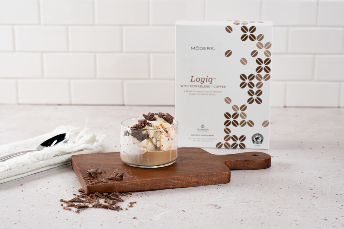 A glass with Modere Logiq Affogato rests on a small, wooden charcuterie board on a kitchen counter. Chocolate shavings spill off one end of the charcuterie board. To the left there is a spoon resting on a folded dish towel. To the right is a box of Modere Logiq.