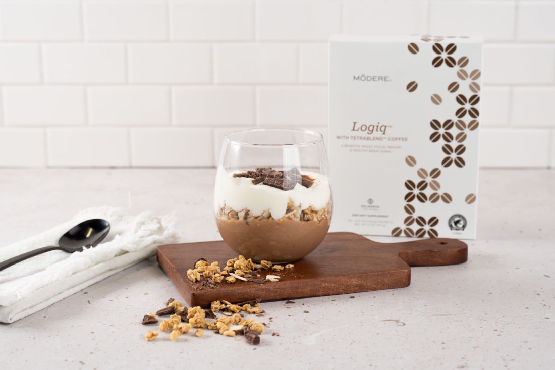 A glass holding Modere Logiq Tiramisu Yogurt Parfait rests on a small, wooden charcuterie board on a kitchen countertop. Granola spills off the charcuterie board. To the left a small spoon rests on a folded dish towel. To the right is a box of Modere Logiq.