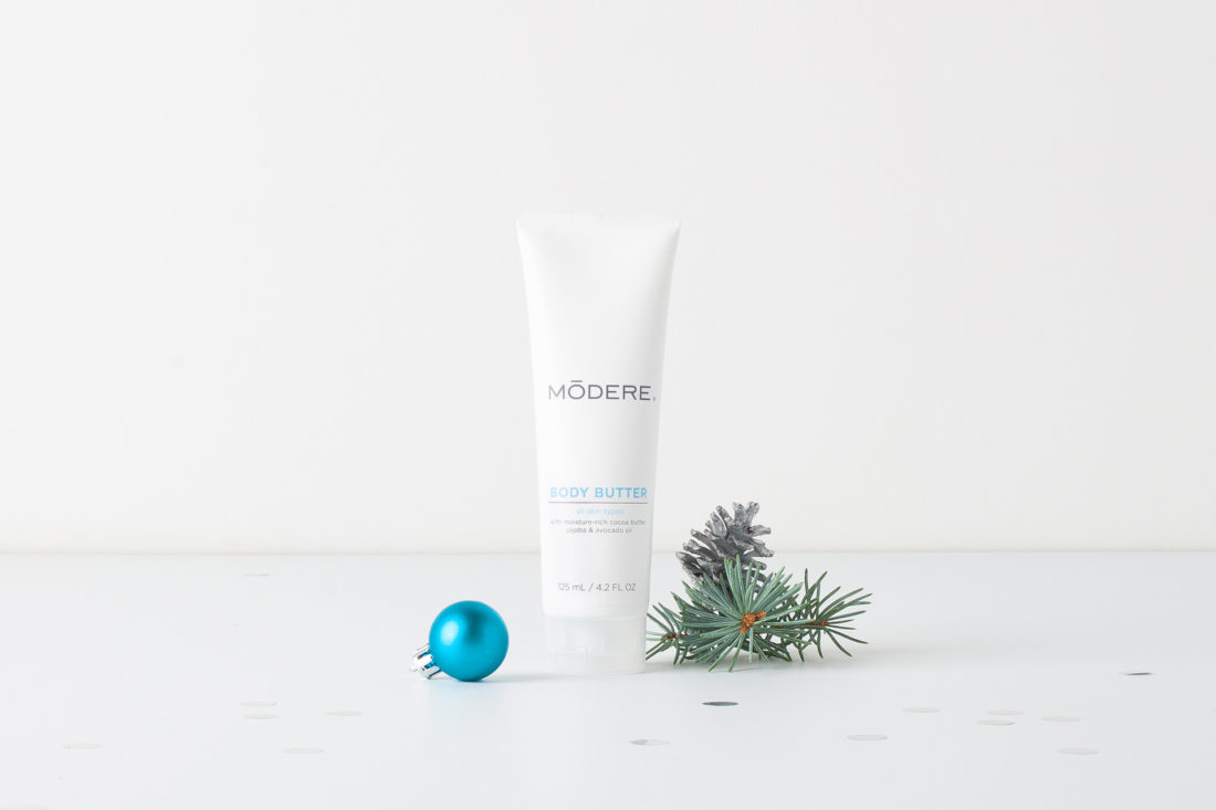 A tube of Modere Body Butter. On either side of the tube are a blue Christmas ornament and a small clipping of a branch from a Christmas tree.