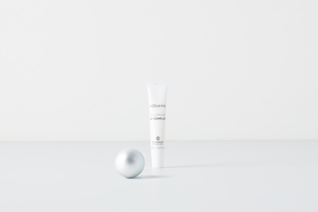 A tube of Modere CellProof Lip Complex. Next to the tube is a small, silver Christmas ornament.