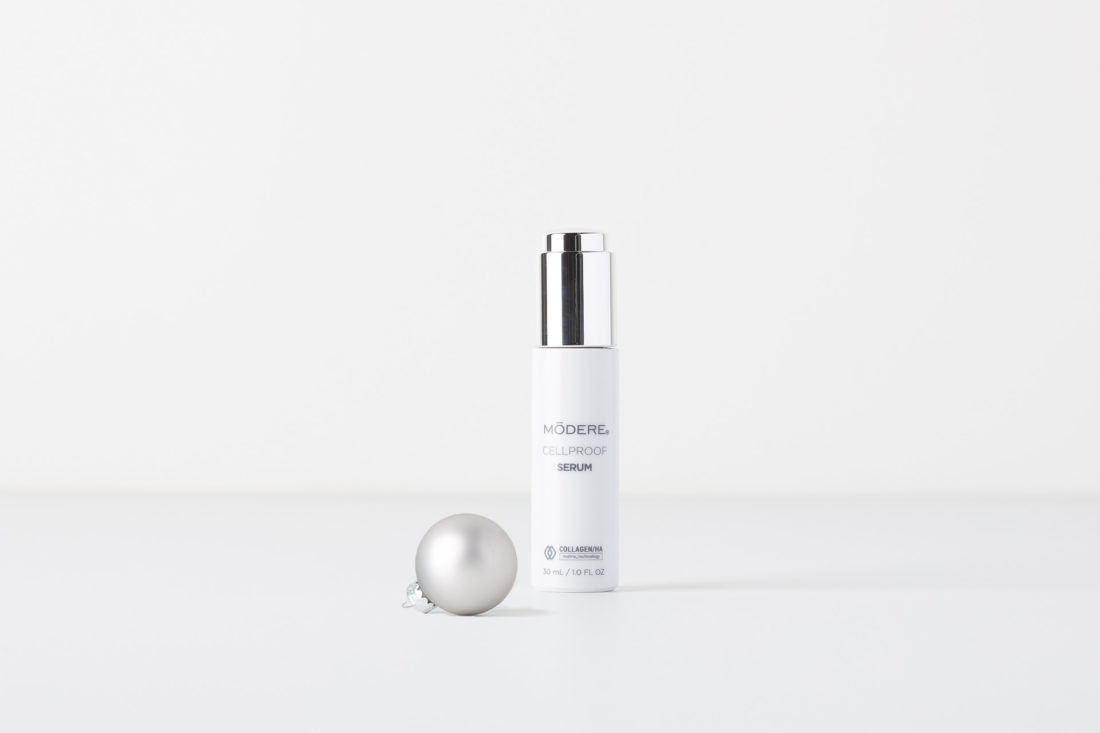 A bottle of Modere CellProof Serum. Next to the bottle is a small, silver Christmas ornament.