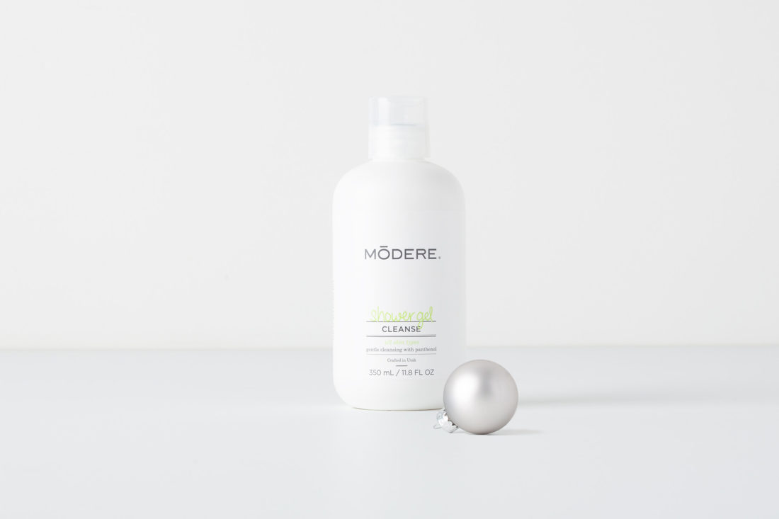 A bottle of Modere Shower Gel on a plain background. There is a silver Christmas ornament in front of the bottle.