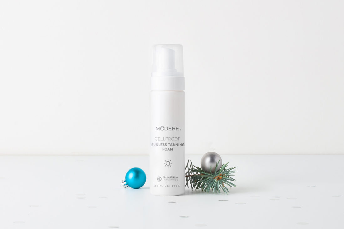 A bottle of Modere CellProof Sunless Tanning Foam. On either side of the bottle are a small blue Christmas ornament, a small silver Christmas ornament, and a small clipping from the branch of a Christmas tree.