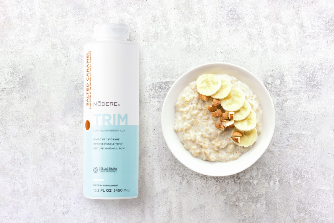A bottle of Modere Trim Salted Caramel next to a bowl of oatmeal with banana slices and almonds.