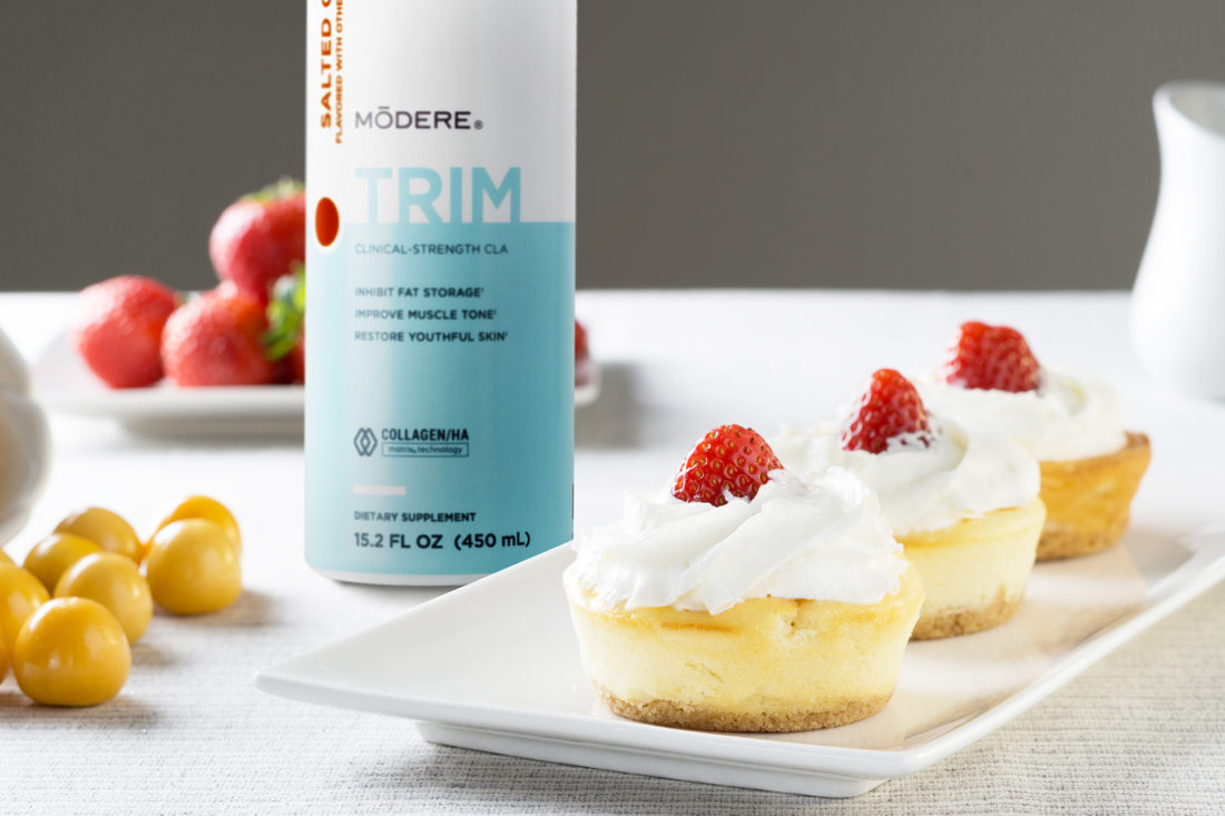 A bottle of Modere Trim Salted Caramel. Next to the bottle is a small plate with three cakes. The cakes are topped with whipped cream and strawberry slices. Behind the bottle of Modere Trim, there are uncut strawberries, and to the left of the bottle is a small bunch of cherries.