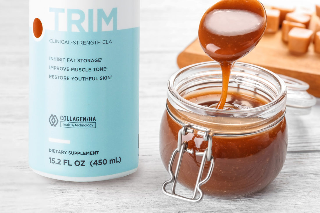 A bottle of Modere Trim Salted Caramel next to a jar of caramel sauce. Behind the caramel sauce we see a wooden cutting board with soft caramels on it.