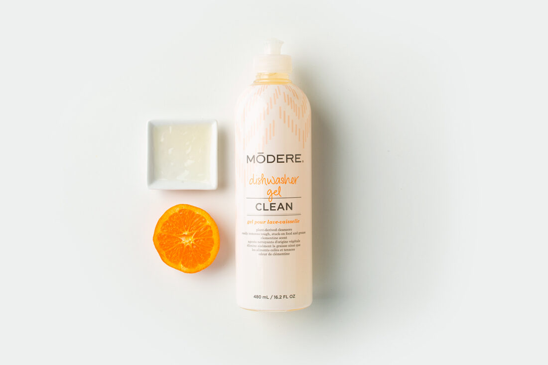 Flat lay image of a bottle of Modere Dishwasher Gel, an orange slice, and a small dish full of Modere Dishwasher Gel on a plain white background.