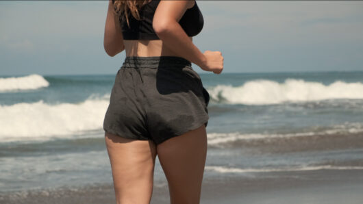 Cellulite is visible on a woman's leg as she runs along a beach.